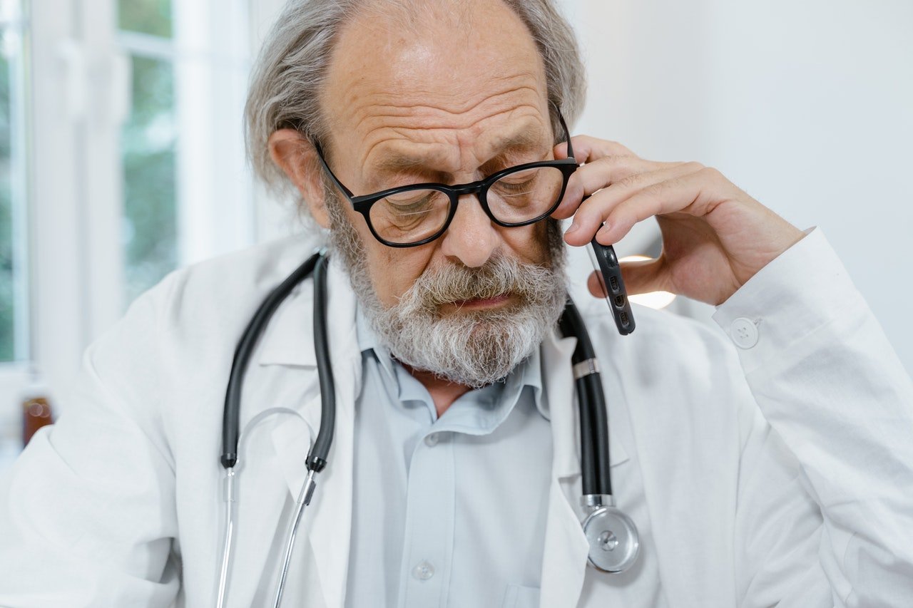 A doctor wearing black glasses and a white jacket speaks on the phone to hire an answering service