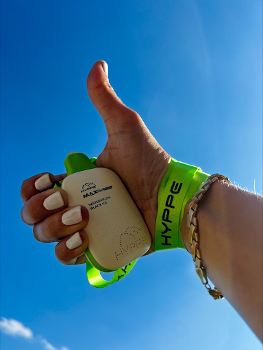 An upward view capturing a hand against a clear blue sky, giving a thumbs up while holding a white, handheld vaping device with a neon green strap. The device has the branding "HYDE" and "MAX AIRFLOW" printed on it, along with the flavor "Watermelon Black Ice" indicated below. The wearer has neatly manicured nails with white polish and wears a simple gold bracelet.