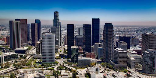 A panoramic view of downtown Los Angeles under clear blue skies, showcasing its dense cluster of high-rise buildings, with the tallest skyscrapers standing prominently against the urban backdrop. The city's sprawling streets and freeways weave through the scene, punctuated by pockets of urban greenery and the hustle and bustle of city life. The horizon fades into a soft haze, highlighting the vast expanse of the city extending into the distance.
