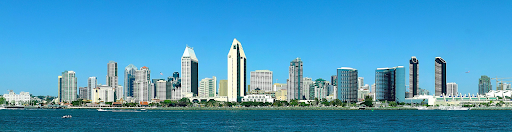 This is a panoramic image of the San Diego skyline viewed from across the water. It features a series of skyscrapers with distinct architectures, including the notable pointed rooftops of the twin towers in the center. The buildings are set against a clear blue sky, with a few small boats visible on the water. A plane can be seen in the distance, flying high above the city. The waterfront is lined with lush trees and smaller structures, contributing to a scenic urban coastline.