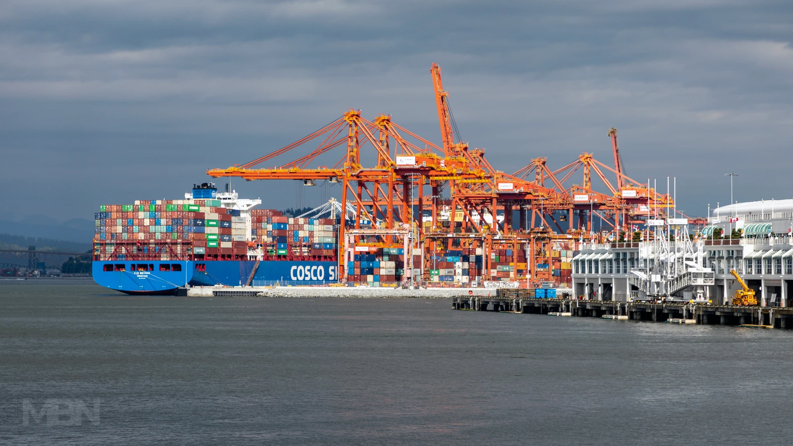 A COSCO container ship moored beside orange cargo cranes at Vancouver Centerm Terminal, with stacked containers and water in the foreground.