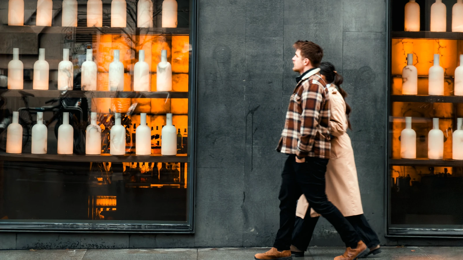 Two pedestrians walk past illuminated shop windows displaying rows of pale bottles beside a grey stone wall in Madrid.