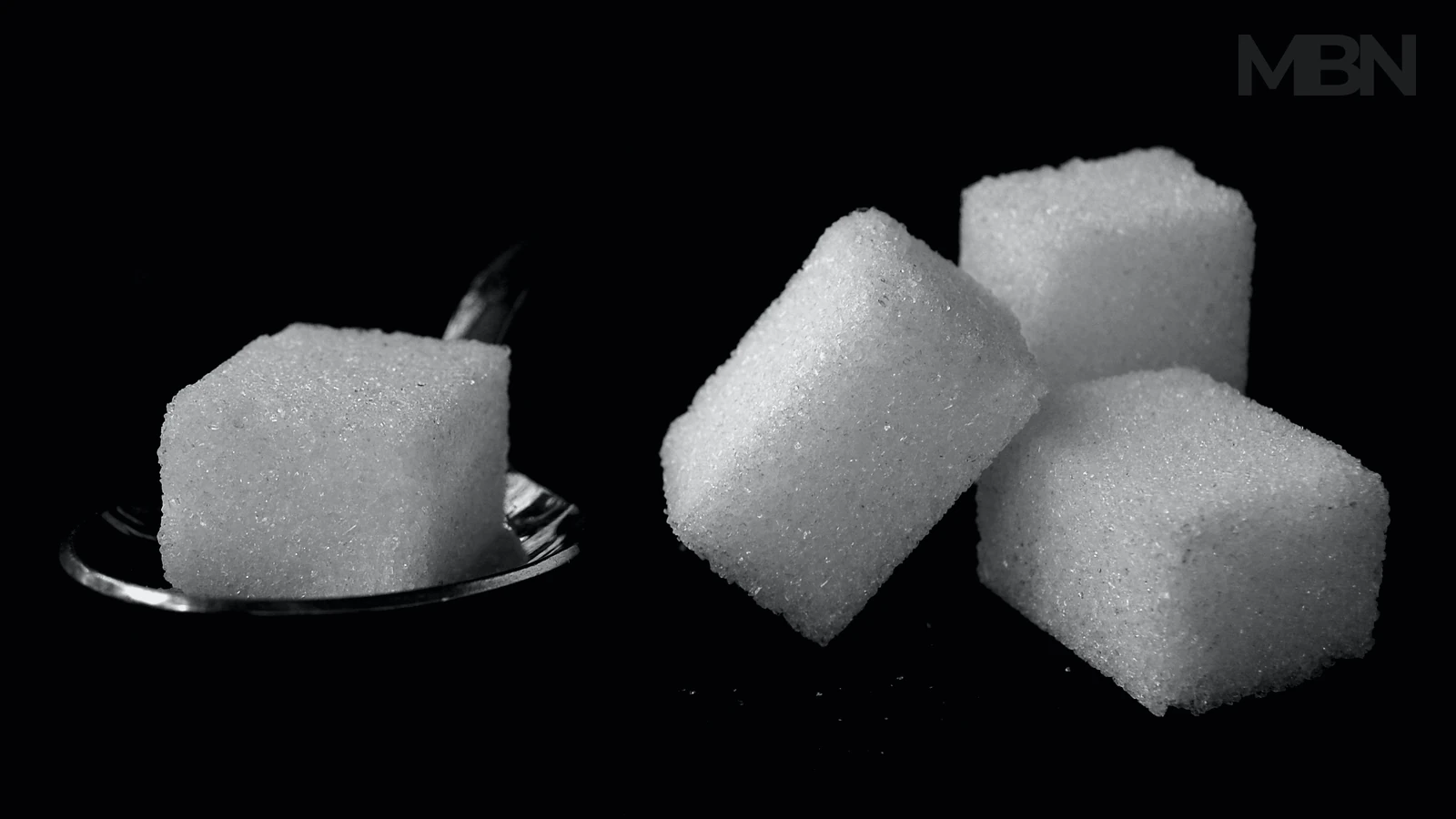 Four white sugar cubes sit against a dark background, with one resting on a metal spoon and a small MBN watermark above.