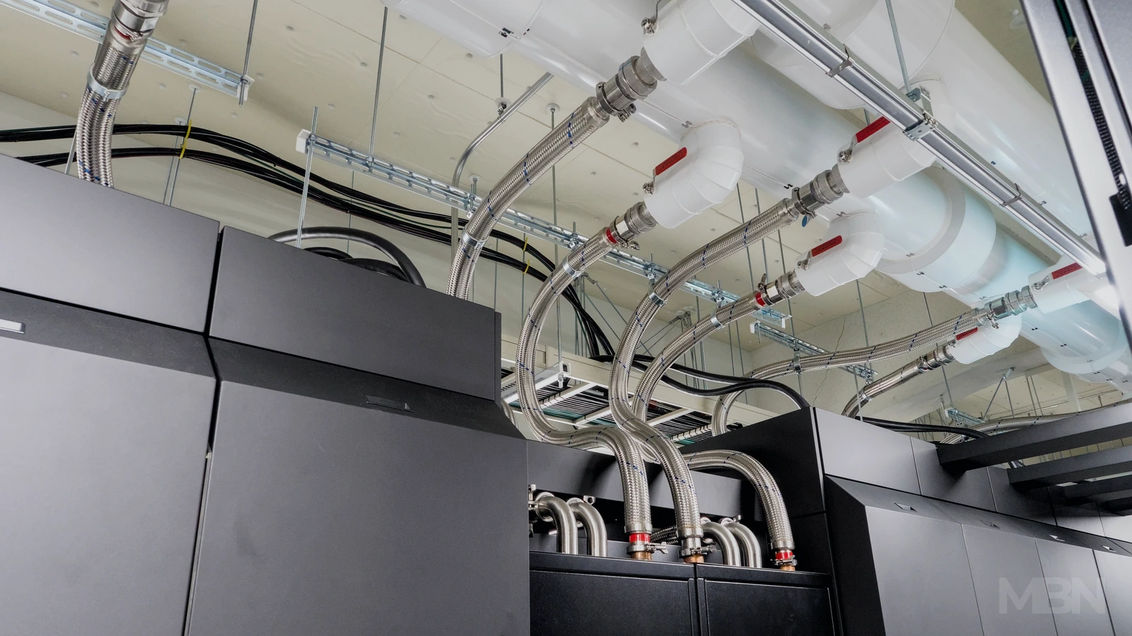 Silver cooling hoses connect overhead white pipes to dark supercomputer cabinets, with black cables running above the equipment.