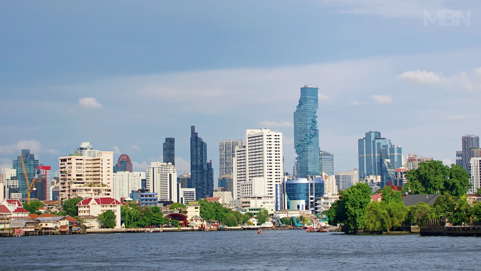 Bangkok’s waterfront skyline, with modern towers and lower riverside buildings above the Chao Phraya River beneath a blue sky.