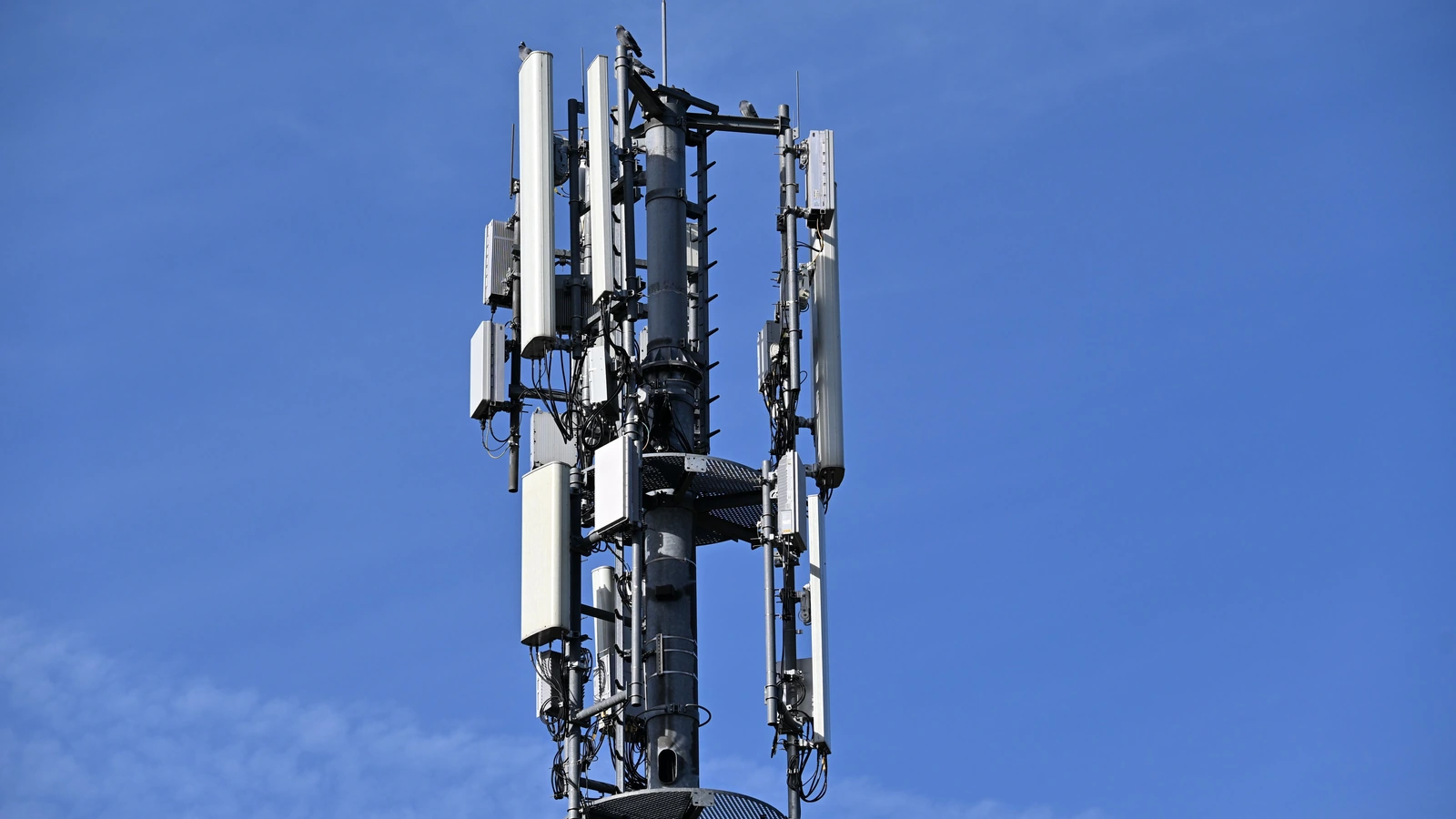 Communications antennas and cables mounted on a mast against a blue sky, representing existing mobile infrastructure; photograph by Edvin Gál/Pexels.