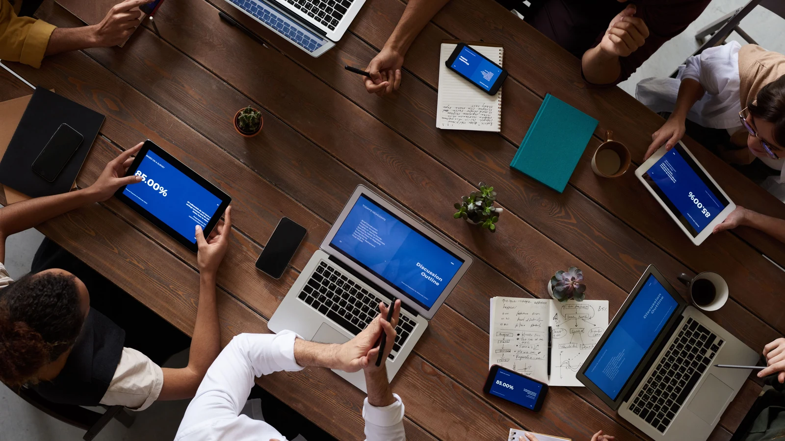 Overhead view of people discussing documents and screens around a wooden table, a representative stock photograph by fauxels/Pexels.