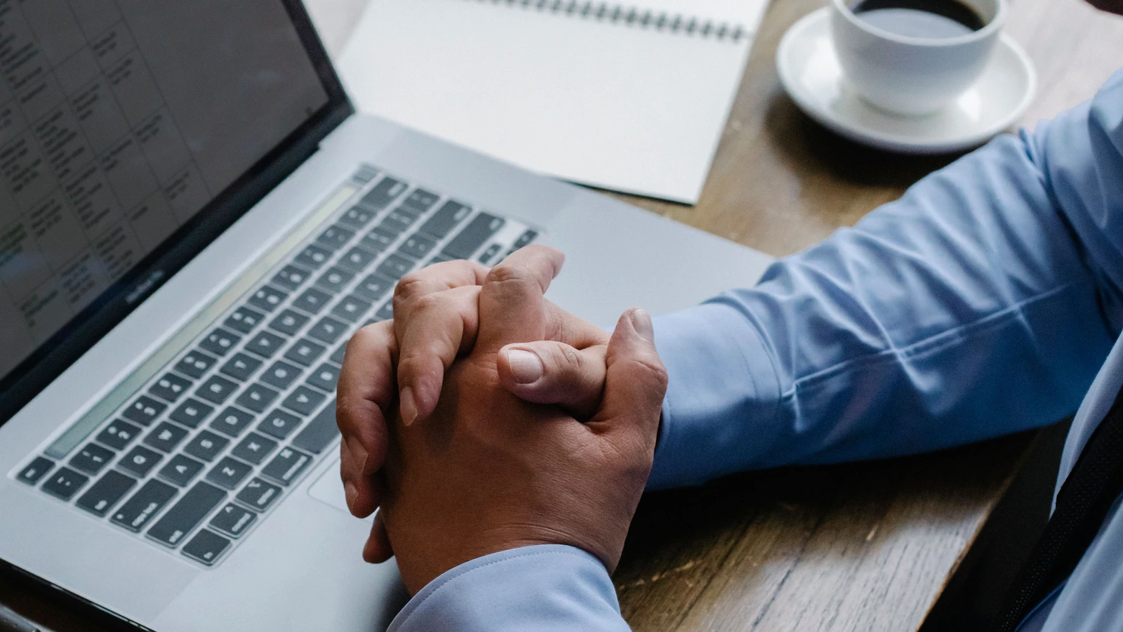 Clasped hands beside a laptop, notebook and coffee, a representative business research photograph by Sora Shimazaki/Pexels.