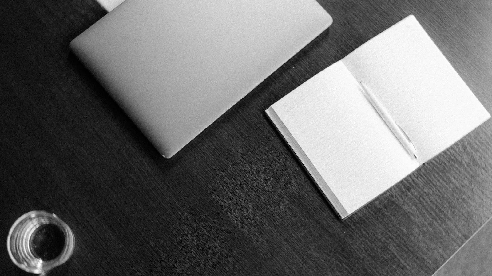 A closed laptop, open notebook, pen and glass on a wooden desk, photographed in black and white by Tima Miroshnichenko/Pexels.