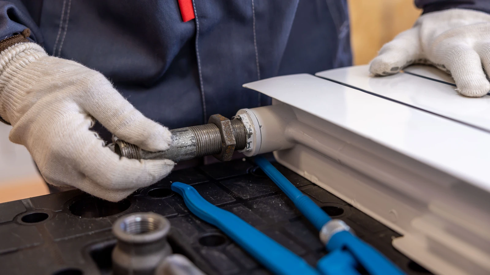 Gloved hands fitting a pipe connection to a radiator, representative stock photography by Sergei Starostin from Pexels under the Pexels License.
