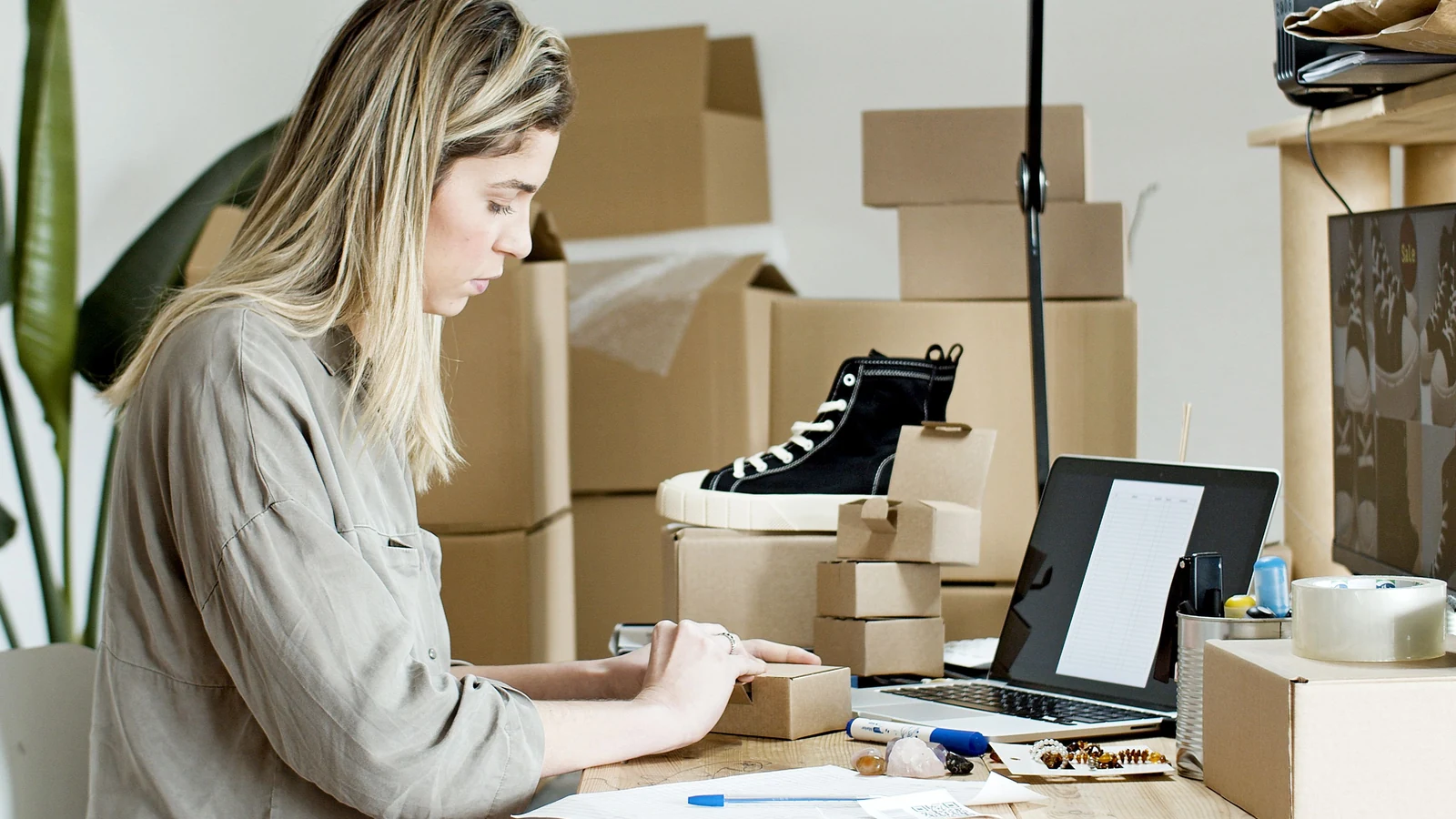 A woman packs a small parcel at a table beside a laptop and boxes. Representative photograph by Kampus Production via Pexels.