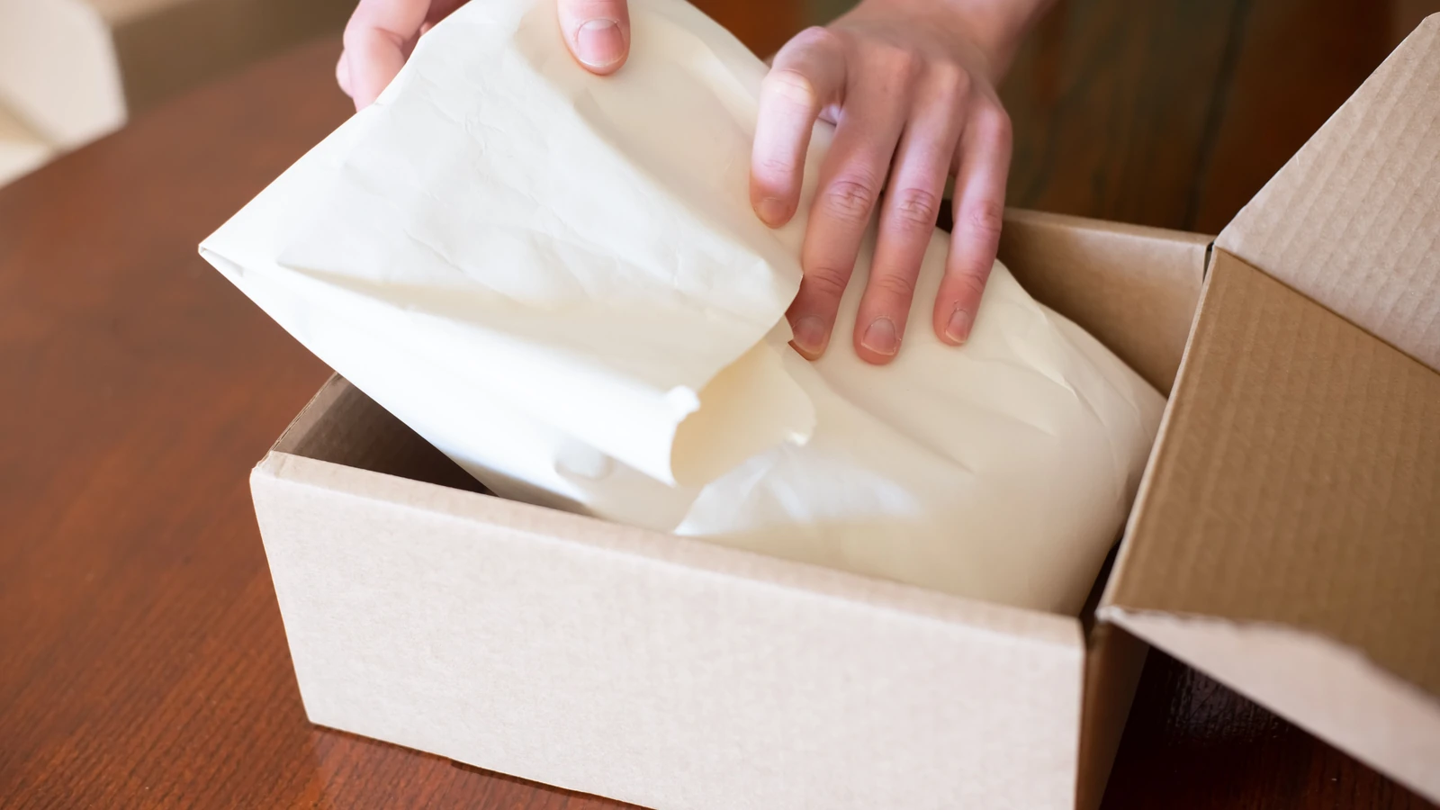 Hands wrapping merchandise in tissue paper inside a cardboard box, representing order fulfillment costs; photograph by Kampus Production/Pexels.