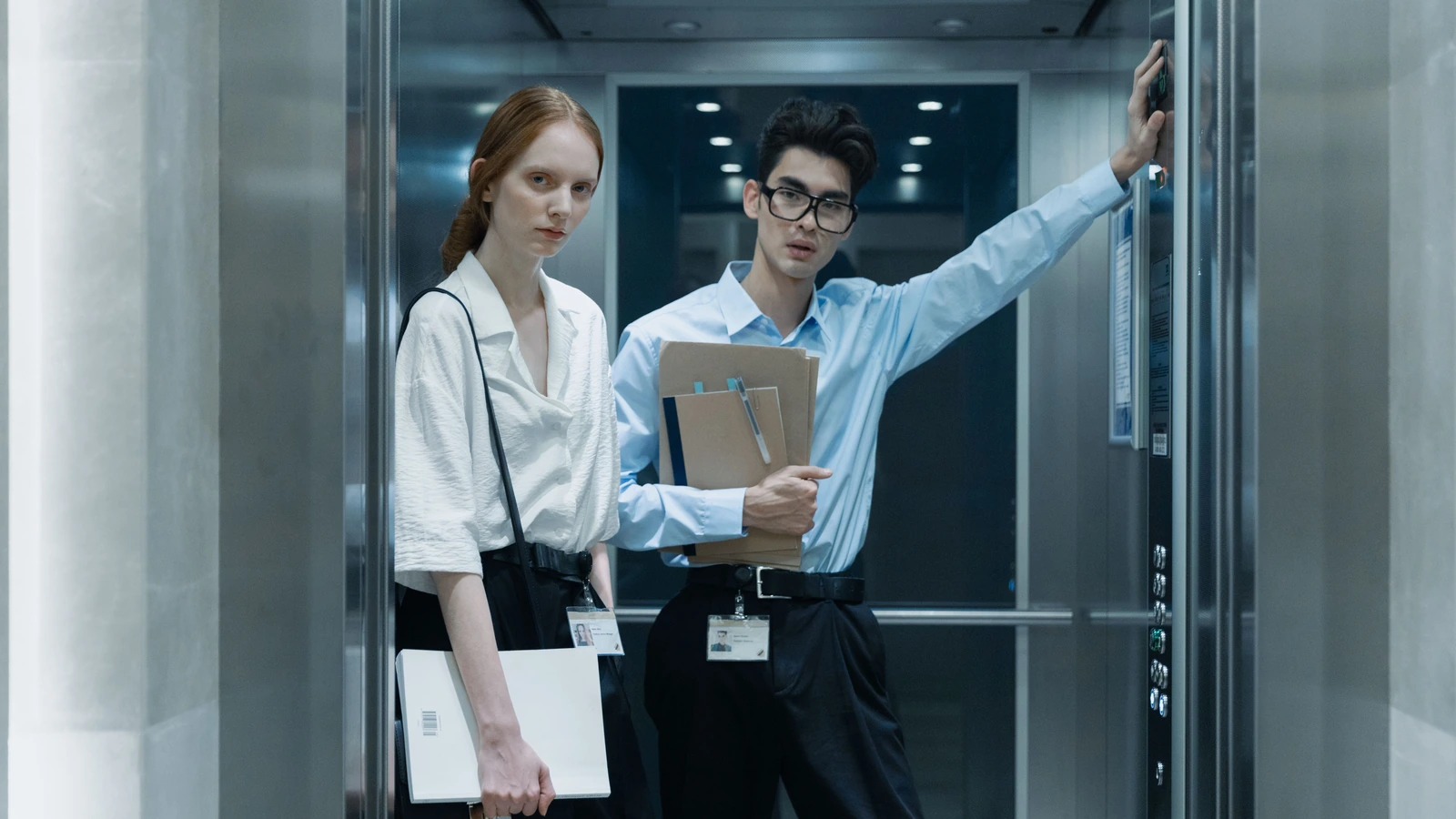 Two people holding folders inside an elevator, one with a hand beside the control panel, in a representative stock photograph by cottonbro studio/Pexels.