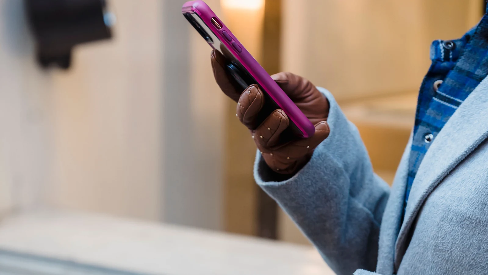 A gloved hand holds a smartphone beside a blue coat in a representative stock photograph by Tim Douglas/Pexels.