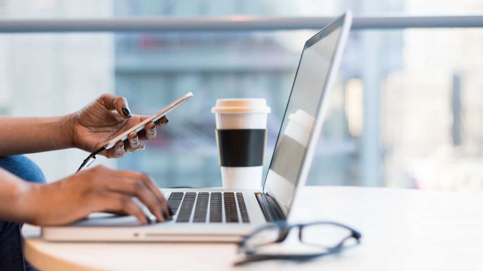 Hands use a laptop and smartphone beside a takeaway coffee, a representative stock photograph by Christina Morillo/Pexels.