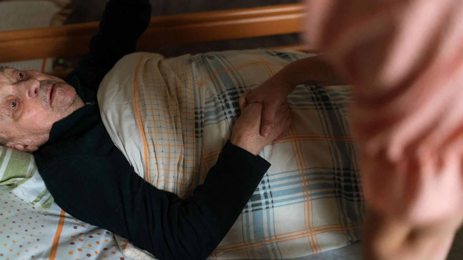 A caregiver holds the hands of an older adult resting in bed, a representative stock photograph by Jsme MILA/Pexels.
