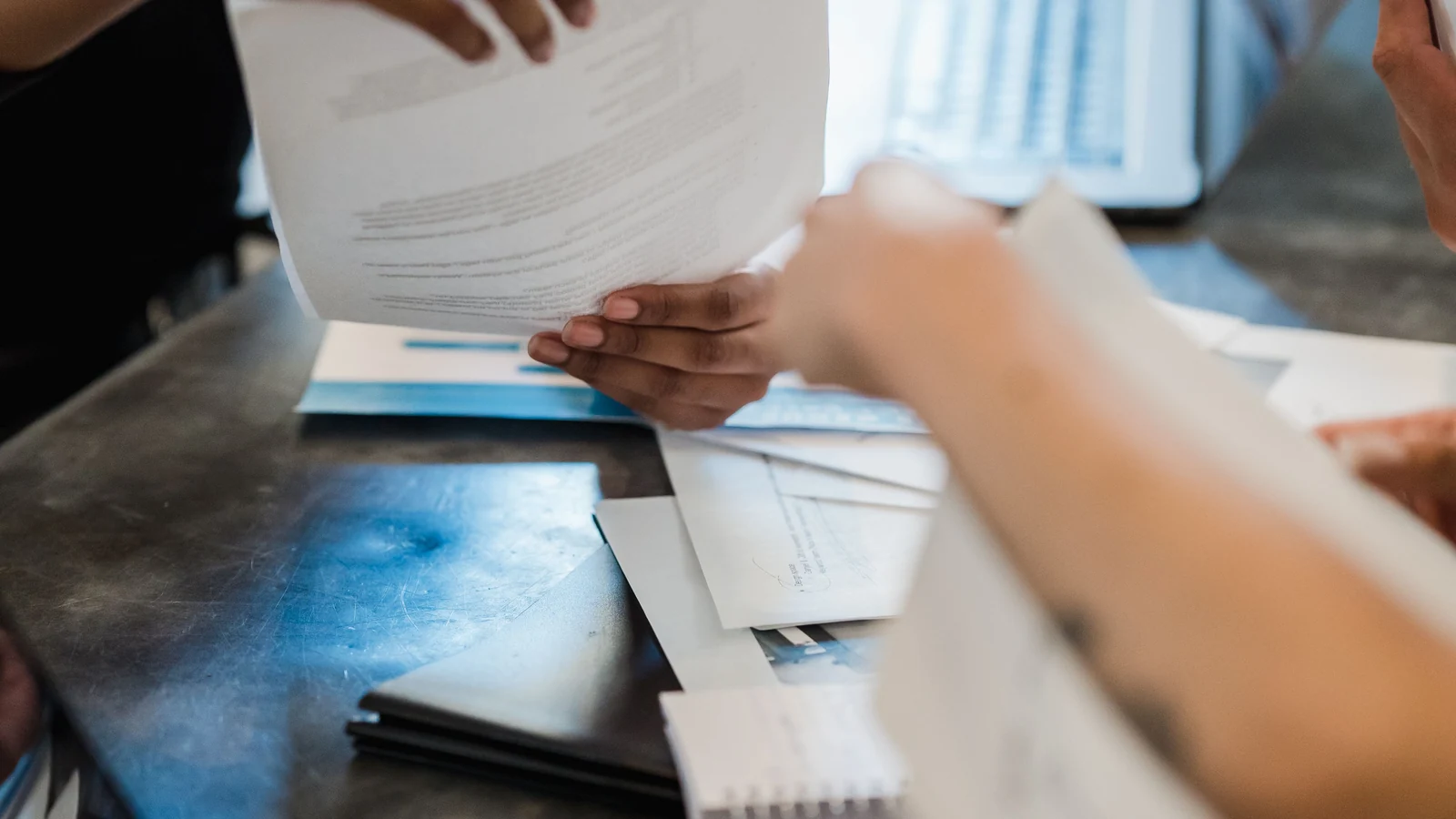 Hands exchange documents across an office table beside a laptop, a representative stock photograph by Mizuno K on Pexels.