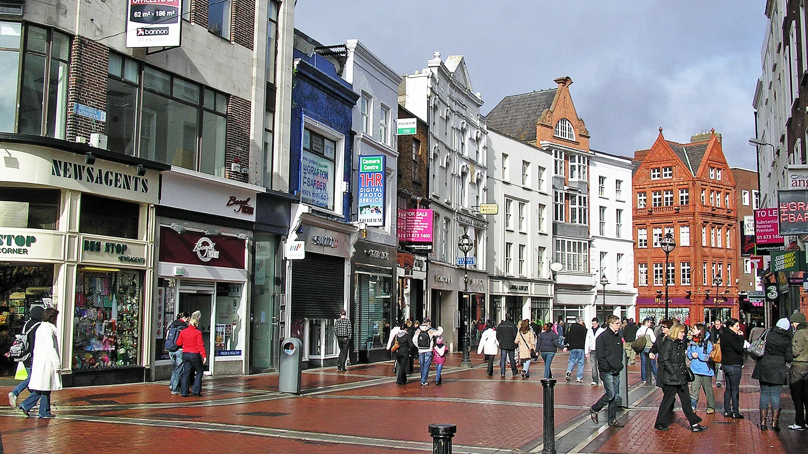 Pedestrians walk past shops and brick-fronted buildings on Grafton Street in Dublin, with a paved pedestrian area in the foreground.