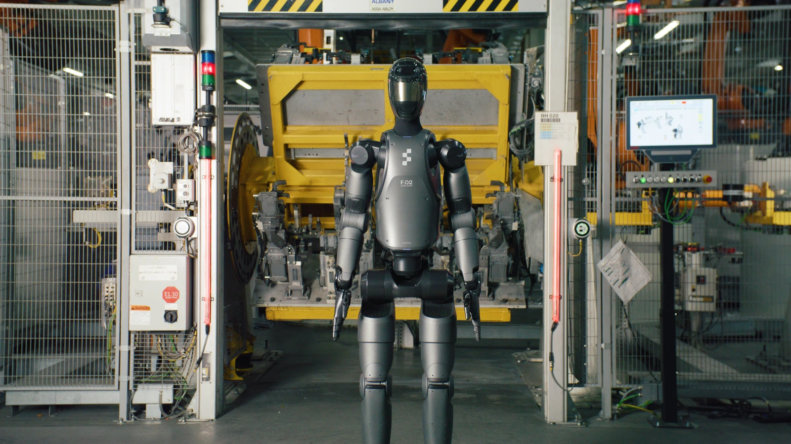 Figure 02 humanoid robot faces the camera inside BMW's Spartanburg factory, with yellow production equipment, safety fencing and a control screen behind it.