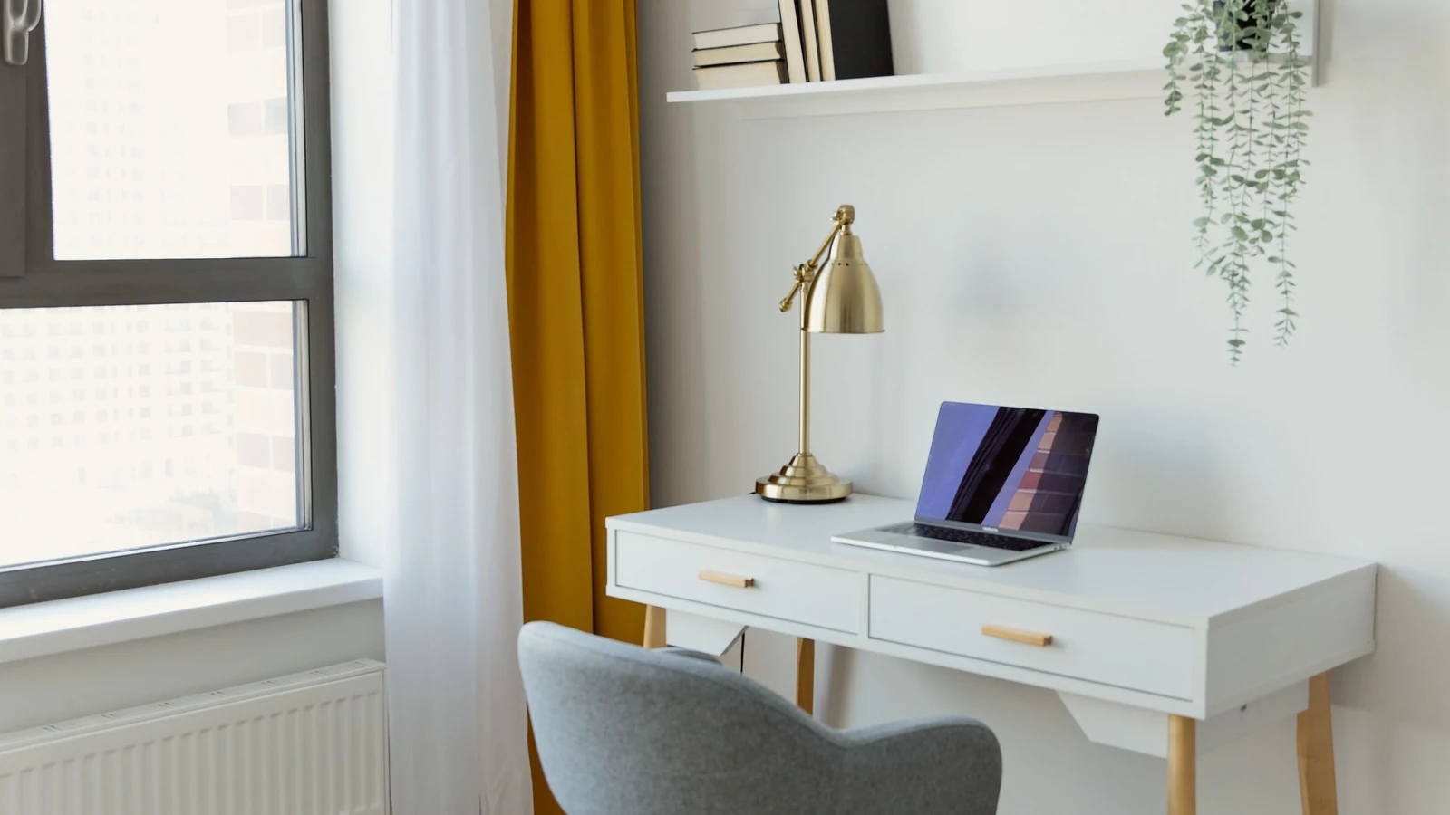 A laptop and lamp on a white desk beside a large window, illustrating a home workspace. Photo by Darina Belonogova/Pexels.