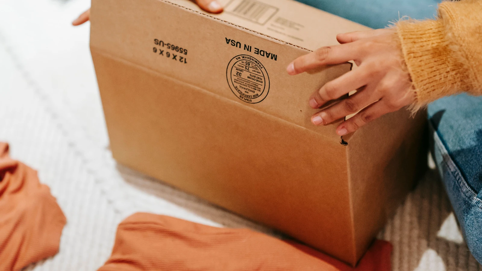 Hands hold a cardboard parcel beside clothing, a representative stock photograph by Liza Summer via Pexels.