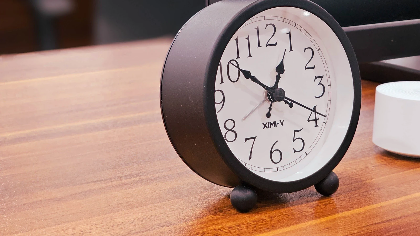 A black analogue clock stands beside a computer monitor on a wooden desk, a representative stock photograph by Zia Moshtaghi/Pexels.