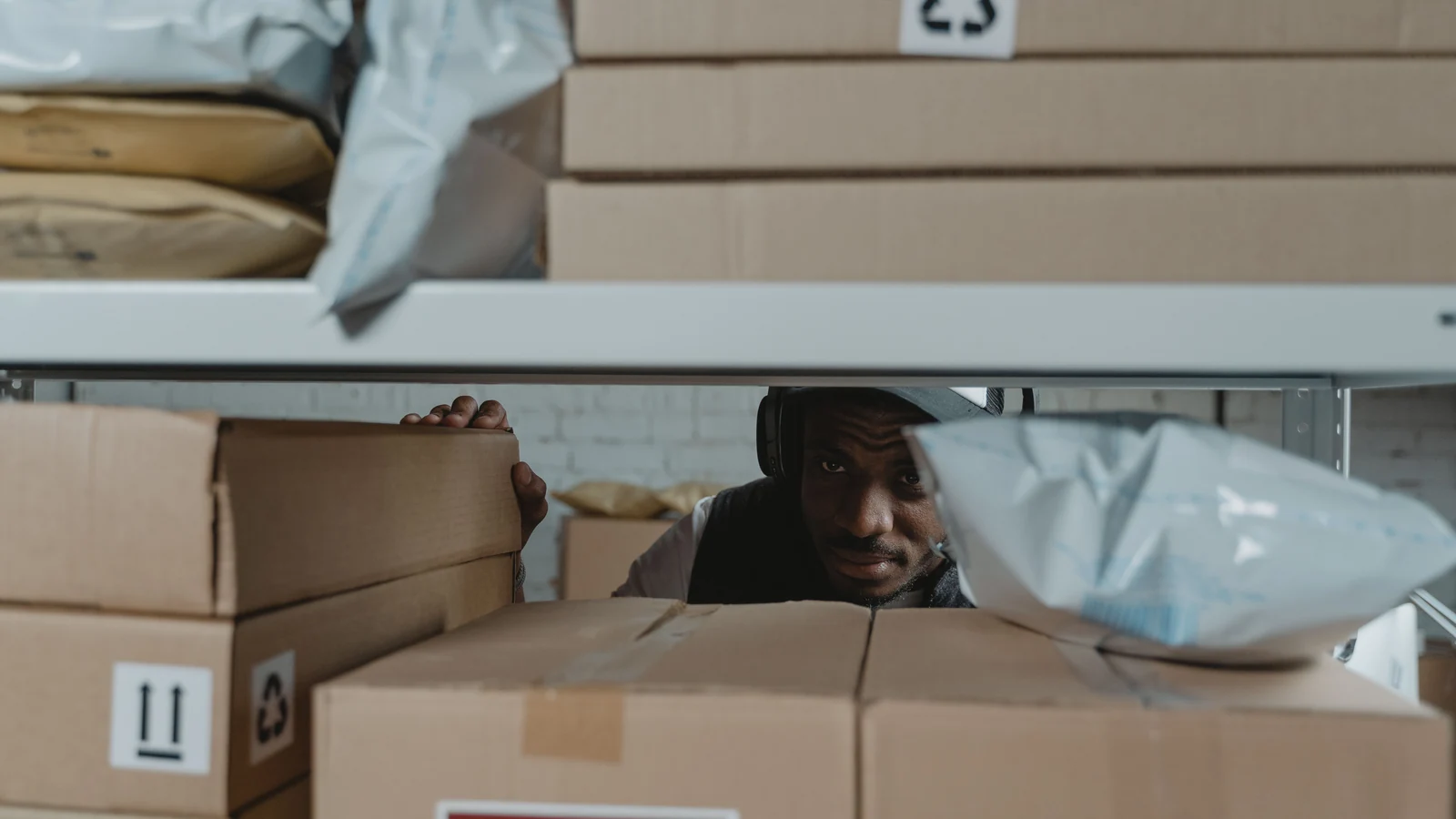 A warehouse worker checks parcels between shelves of cardboard boxes, a representative stock photograph by Tima Miroshnichenko/Pexels.